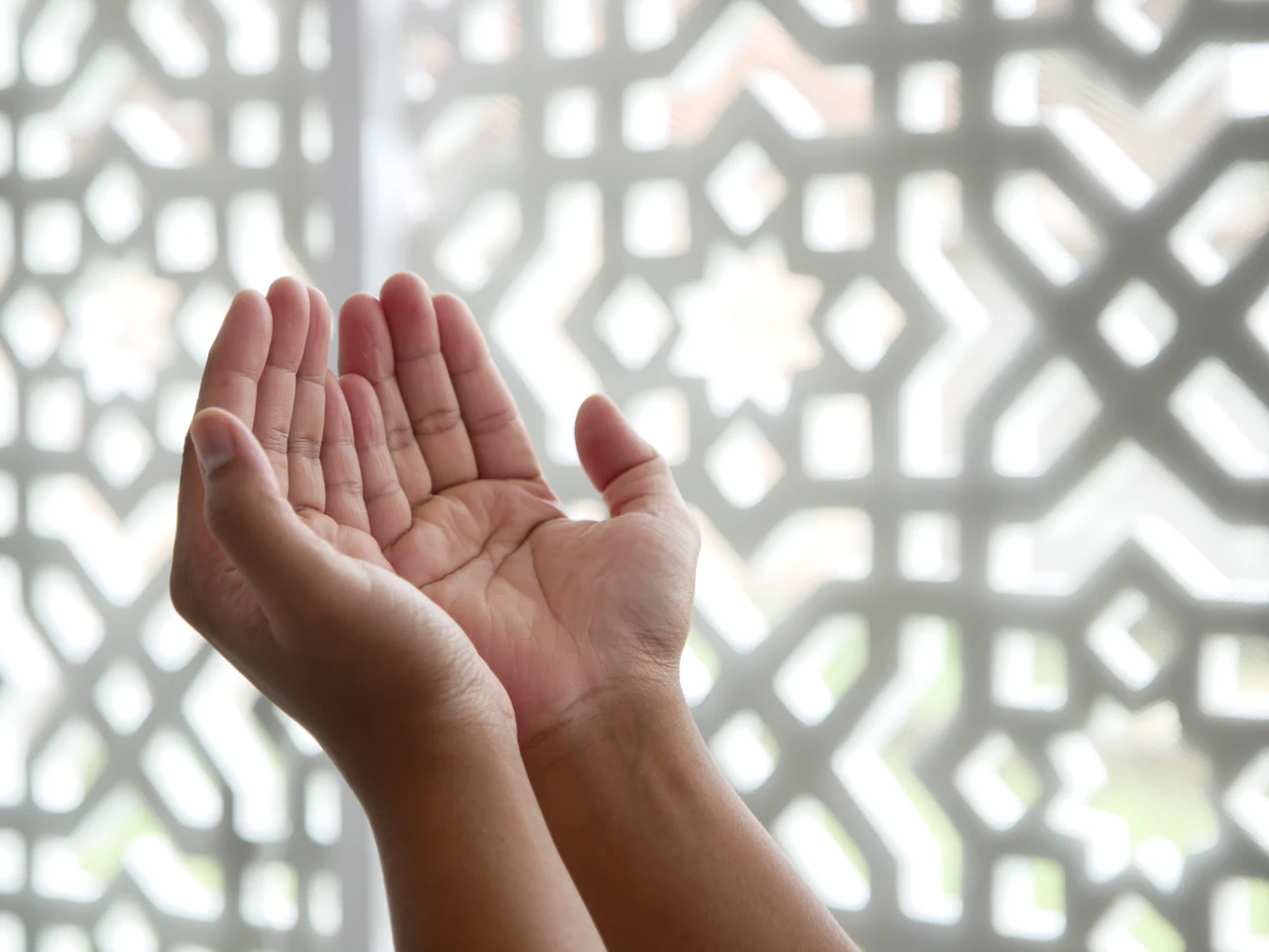 Hands raised in dua in front of a lattice window